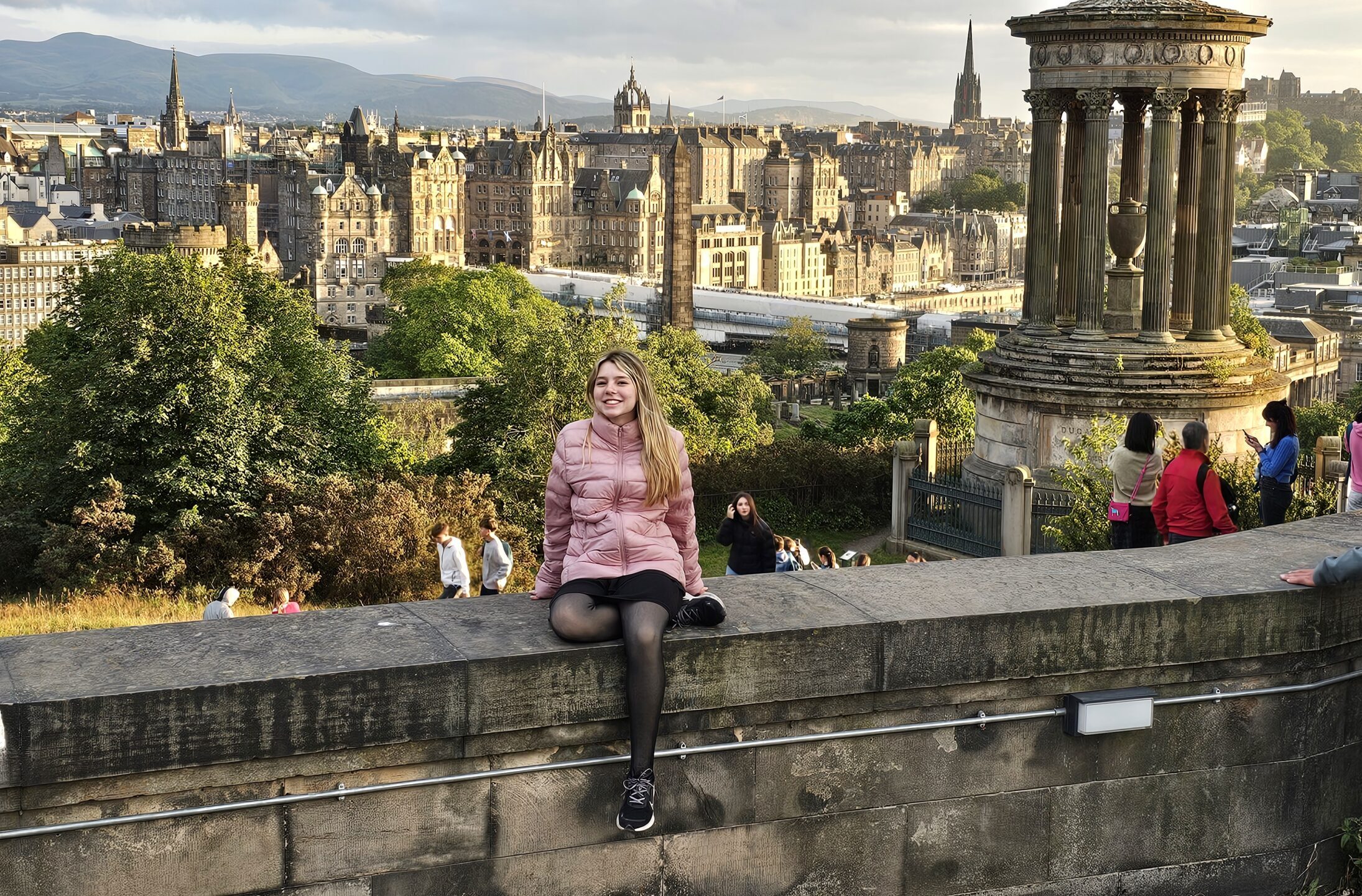 Anna Clara sentada no Calton Hill sorrindo, com o Monumento Dugald Stewart à direita e a Old Town de Edimburgo iluminada pelo pôr do sol ao fundo. Capa do artigo roteiro edimburgo