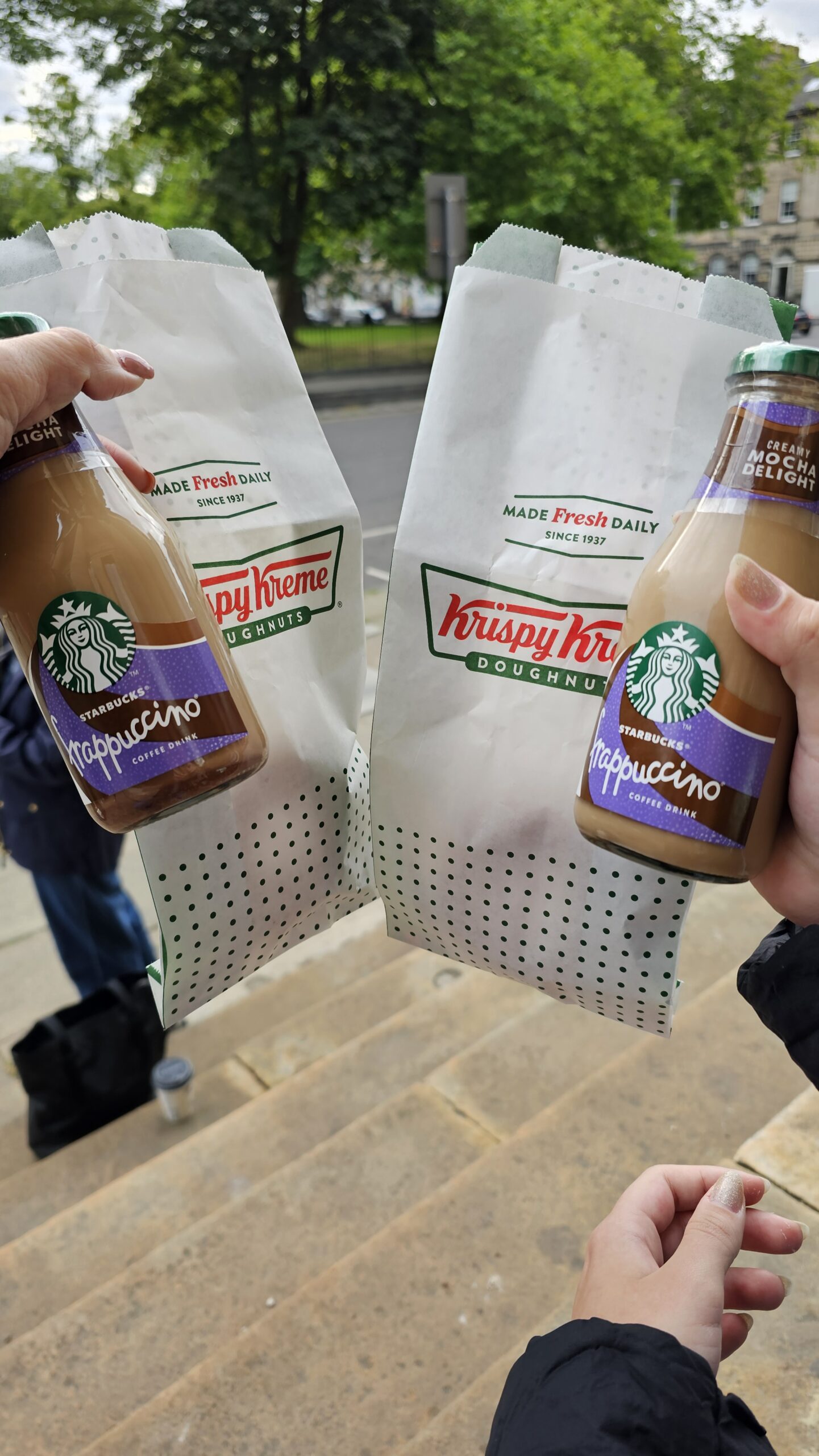 Duas sacolas Krispy Kreme e dois Frappuccinos Starbucks segurados na frente de um parque em Edimburgo, café da manhã antes do tour Highlands.